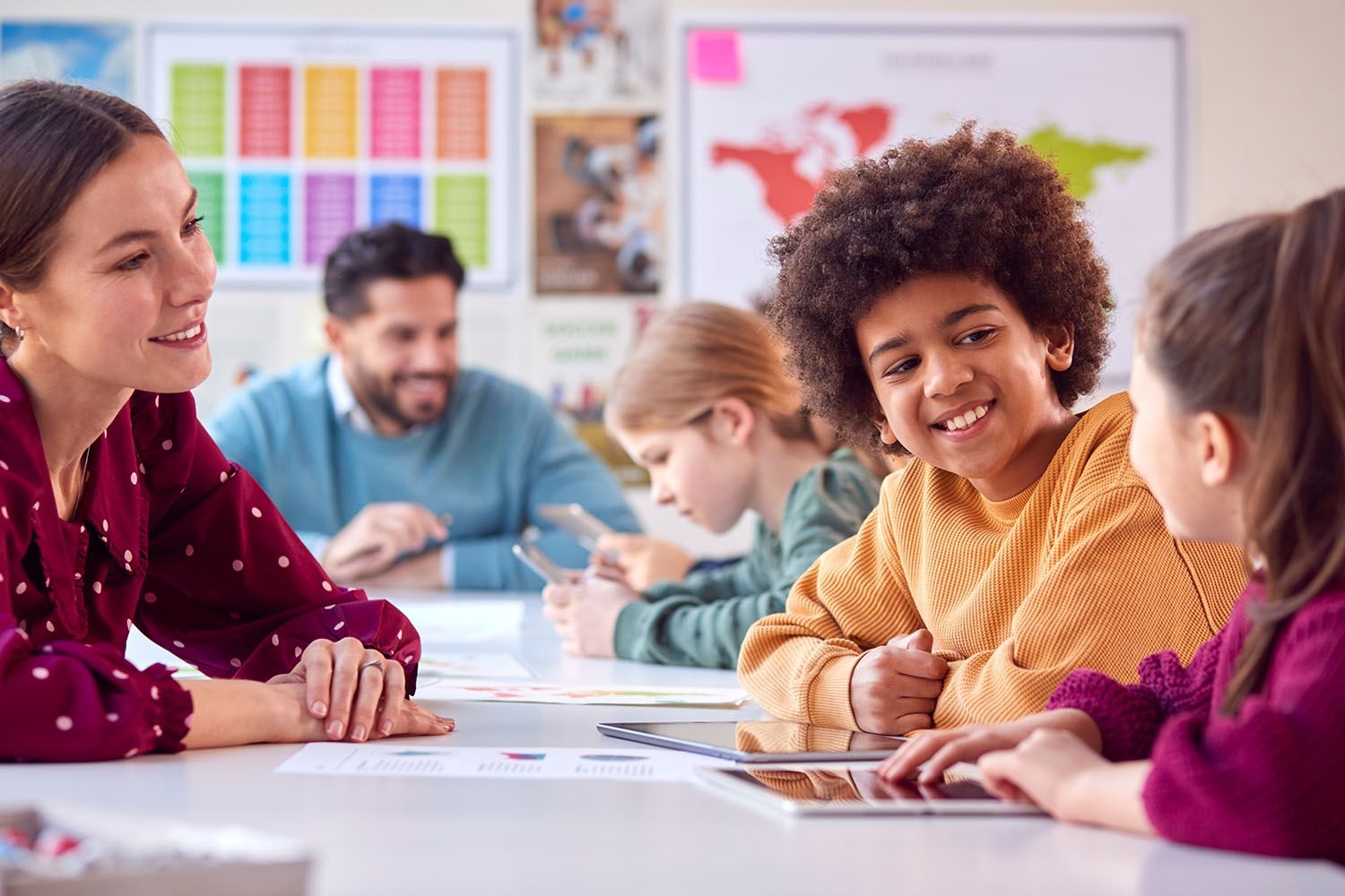 Zwei Lehrkräfte sitzen im Klassenzimmer zusammen mit Schülerinnen und Schülern, die an Tablets arbeiten.